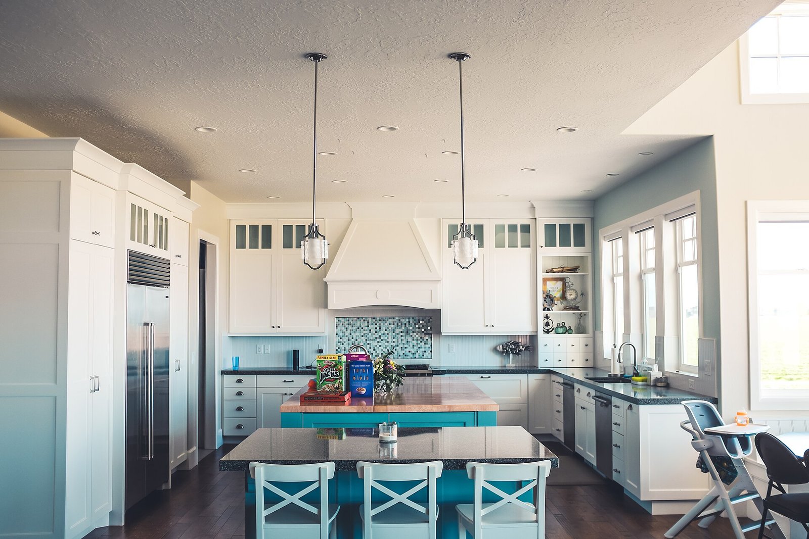 A bright open kitchen with white shaker cabinets, a teal island with butcher-block top, two pendant lights, a granite bar with three stools and a mosaic splashback.