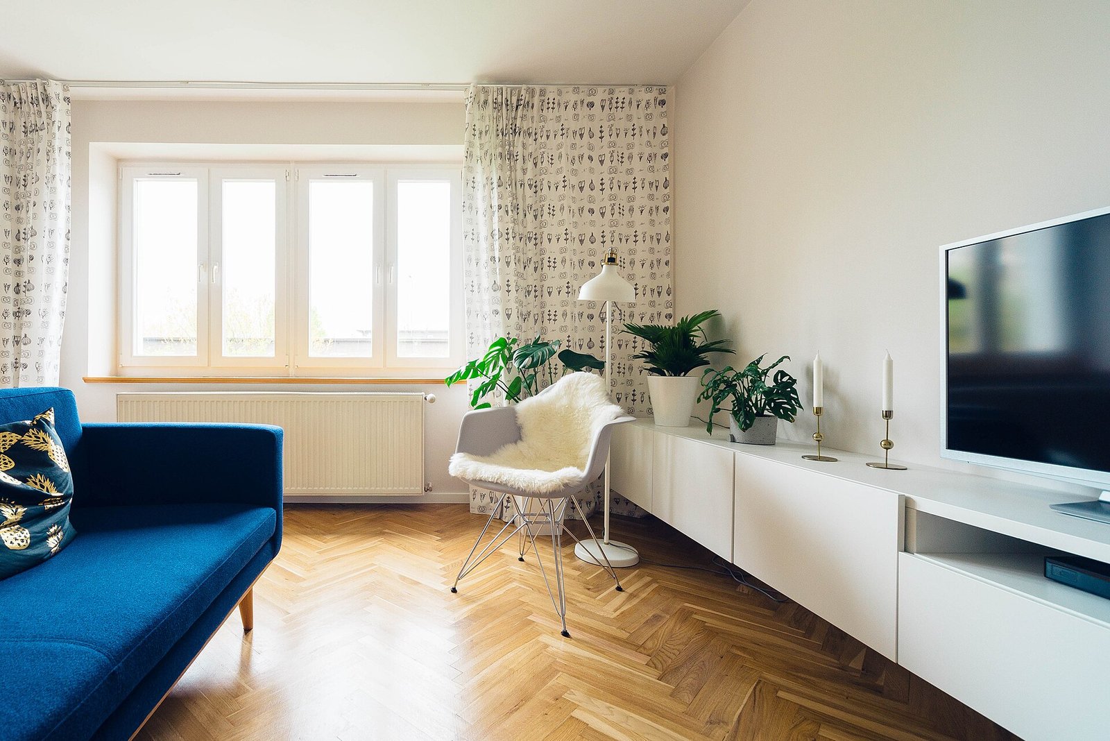 A bright living room with a blue sofa, a white shell chair with a sheepskin, a wall-hung white console with a television, patterned curtains and a radiator under the window.